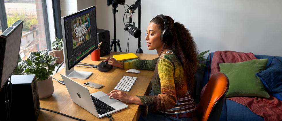 Une femme chez elle, travaillant à son bureau sur un MacBook Pro connecté à deux écrans externes, portant un casque et parlant dans un micro