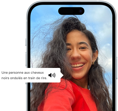 Un iPhone 15 montrant la fonctionnalité VoiceOver énonçant les informations figurant sur l'image : Une personne aux cheveux noirs ondulés en train de rire